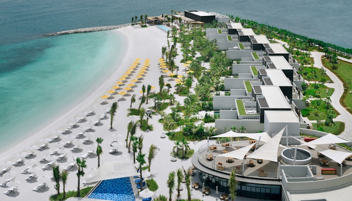 Mövenpick Resort Al Marjan Island Aerial view of a beach resort featuring white sandy beaches, turquoise waters, and modern villas surrounded by lush greenery and lounge chairs.