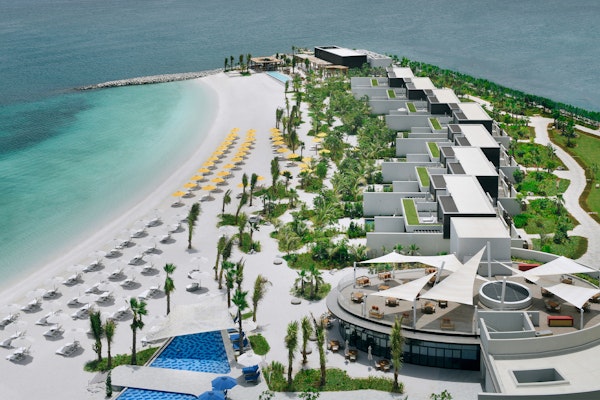 Aerial view of a beach resort featuring white sandy beaches, turquoise waters, and modern villas surrounded by lush greenery and lounge chairs.