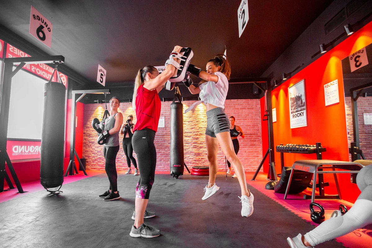 Final Round A group of women engaged in a boxing workout in a gym, with one woman jumping to punch pads held by another.
