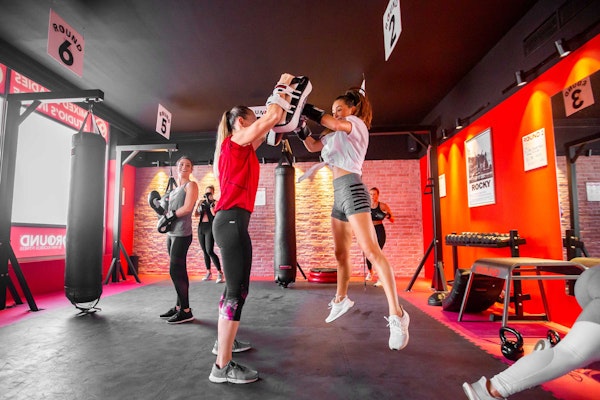 A group of women engaged in a boxing workout in a gym, with one woman jumping to punch pads held by another.