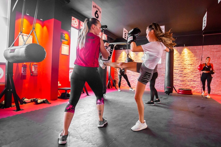 Two women practice kickboxing in a gym, with one delivering a kick to a punching bag held by the other, surrounded by fitness equipment and other gym-goers.