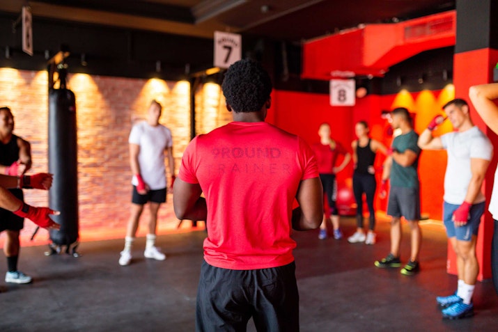 A fitness instructor in a red shirt leads a group of individuals in a boxing workout, with participants gathered around in a gym setting.