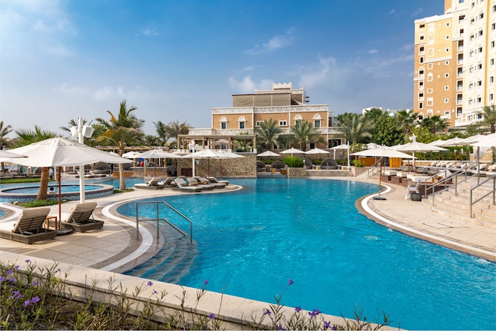 A scenic view of a hotel pool area with sun loungers, umbrellas, and palm trees under a blue sky.