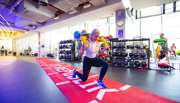 FitRepublik A woman performing a lunge exercise with weights in a modern gym, featuring colorful workout equipment in the background.