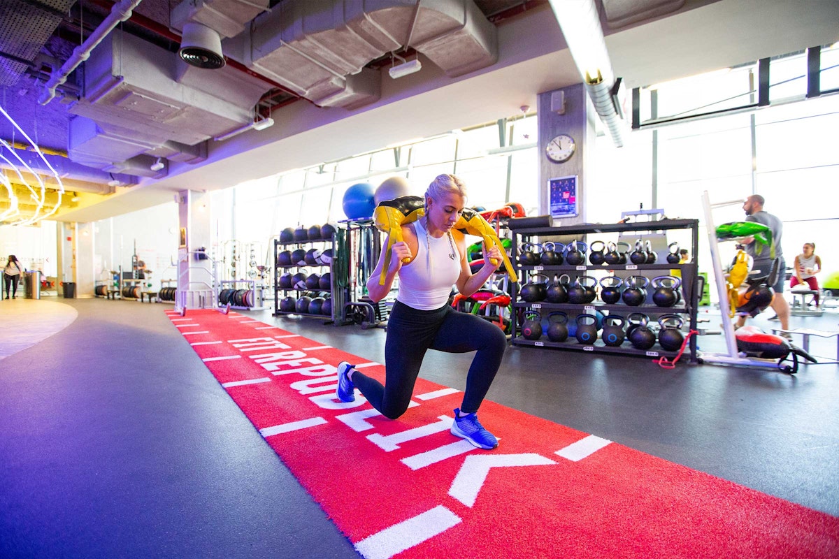 A woman performing a lunge exercise with weights in a modern gym, featuring colorful workout equipment in the background.