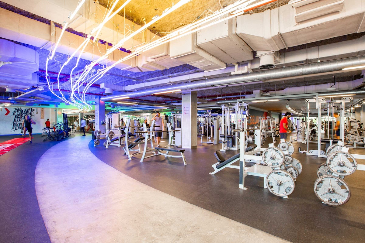 A spacious gym interior featuring various weightlifting equipment and colorful LED lighting, with individuals exercising in the background.