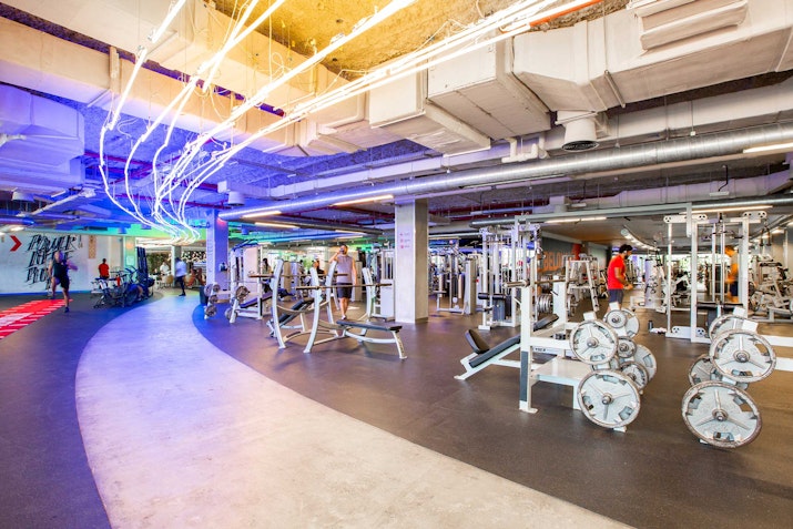 A spacious gym interior featuring various weightlifting equipment and colorful LED lighting, with individuals exercising in the background.