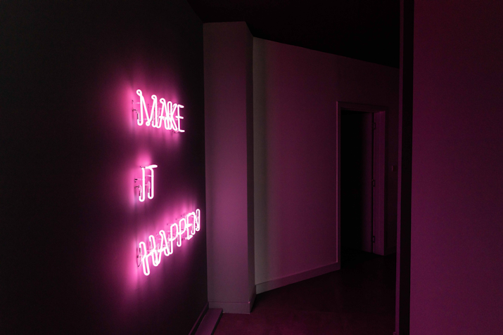 A pink neon sign on a dark wall reading 'MAKE IT HAPPEN,' illuminating a dimly lit hallway with a vibrant glow. The neon light creates an inspiring and motivational ambiance, urging action and positivity
