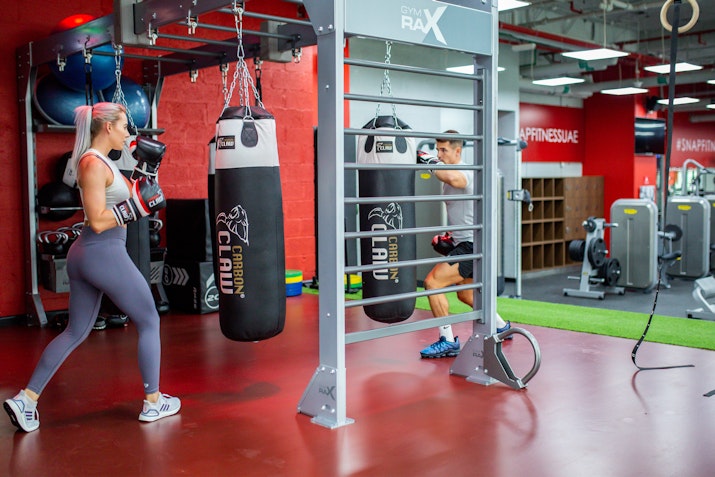A woman practices boxing on a punching bag while a man performs exercises on a nearby fitness station in a vibrant gym.
