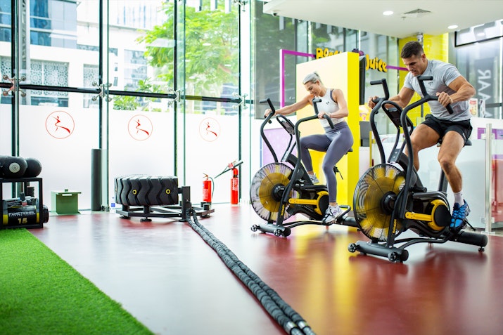 A man and woman workout on stationary bikes in a gym with large windows, surrounded by fitness equipment.