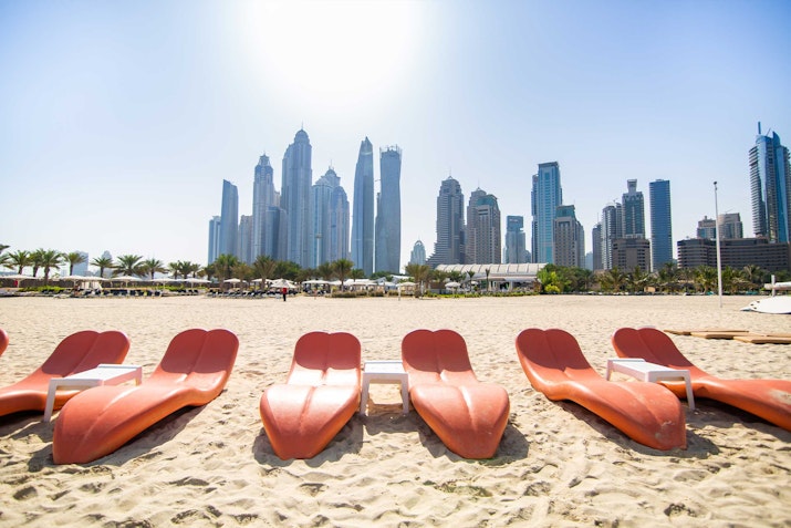 Row of red lounge chairs on a sandy beach with a city skyline in the background.