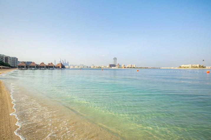Calm beach shoreline with buildings and clear water.