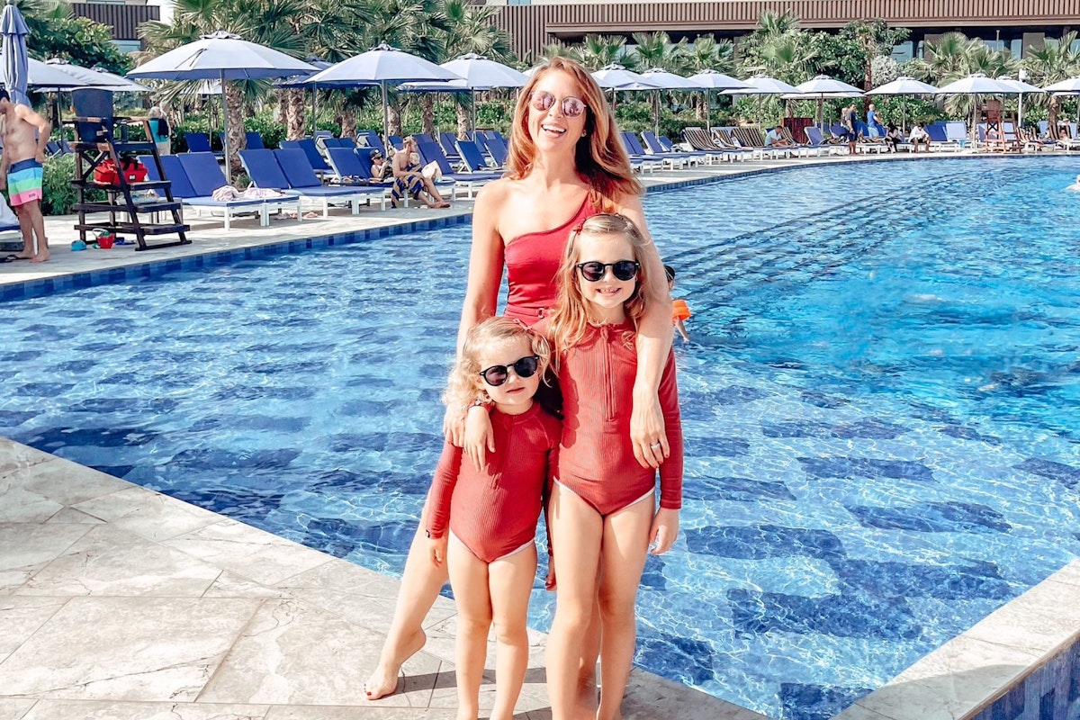 A woman and two children wearing matching red swimsuits and sunglasses, standing by a swimming pool with lounge chairs and umbrellas in the background.