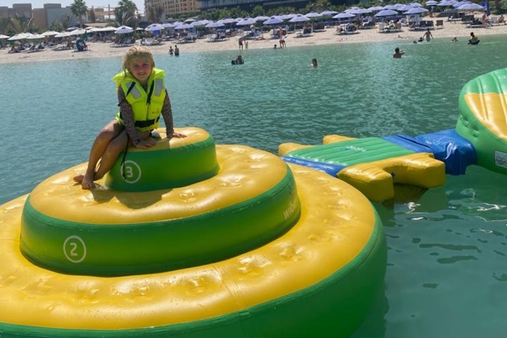 A child in a life jacket sitting on a large inflatable water slide in a pool, with beachgoers and umbrellas visible in the background.