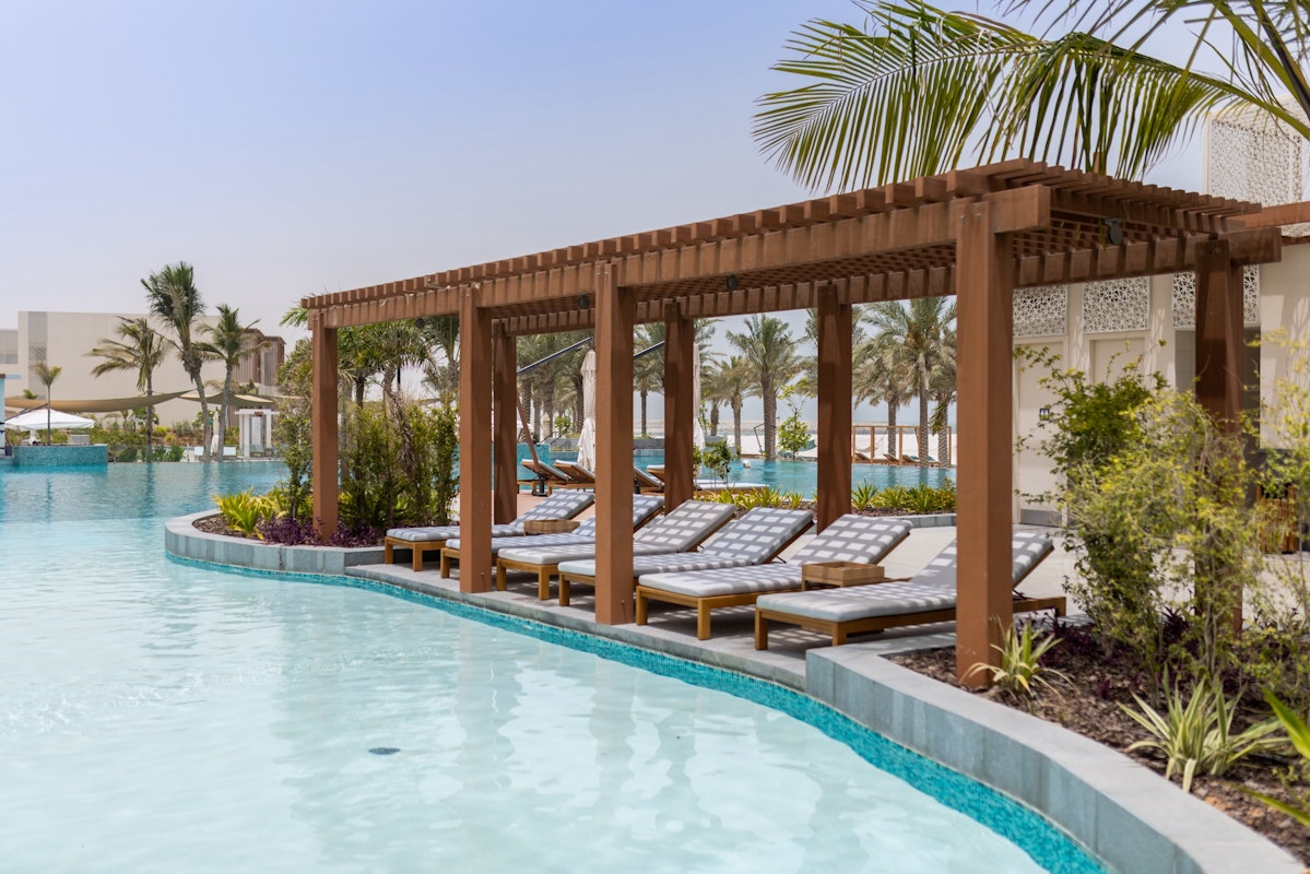 A tranquil pool area featuring a wooden pergola with loungers, surrounded by lush greenery and palm trees.