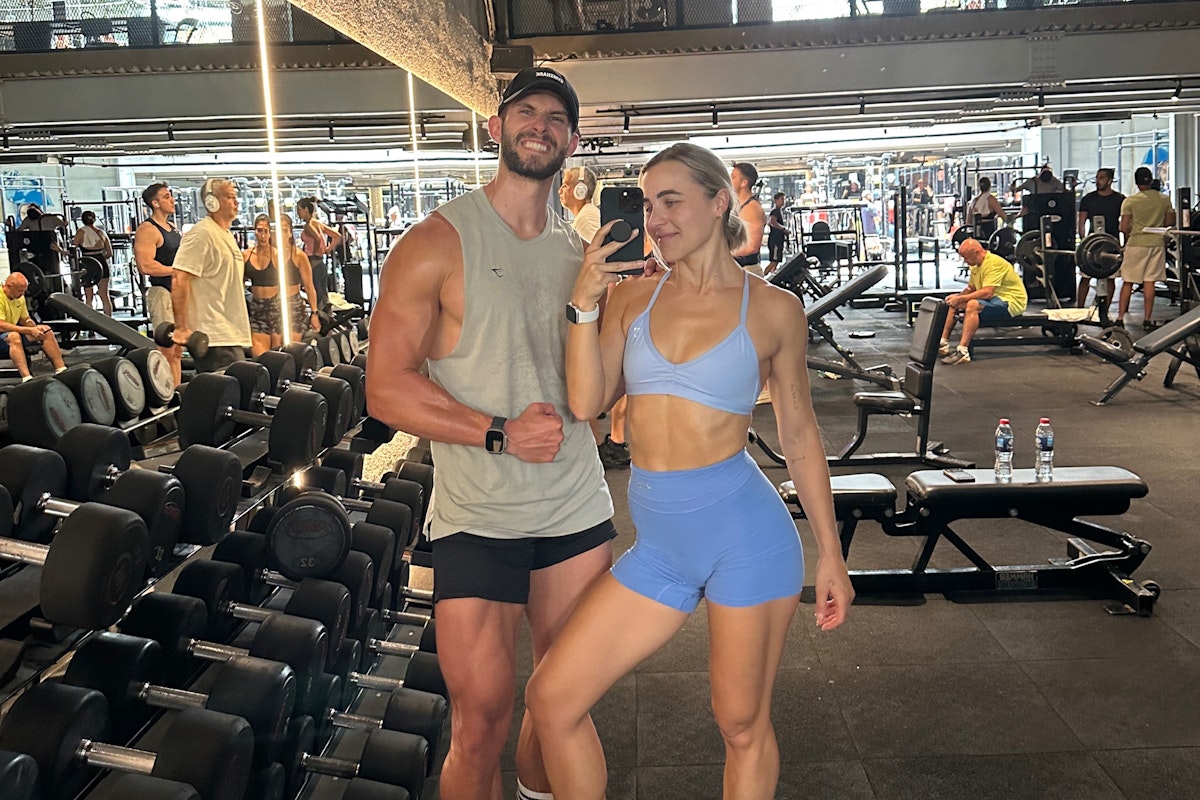 Two people posing for a selfie in a gym with dumbbells in the background.