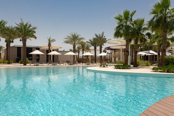 Swimming pool surrounded by umbrellas and palm trees with a sunny sky in the background.