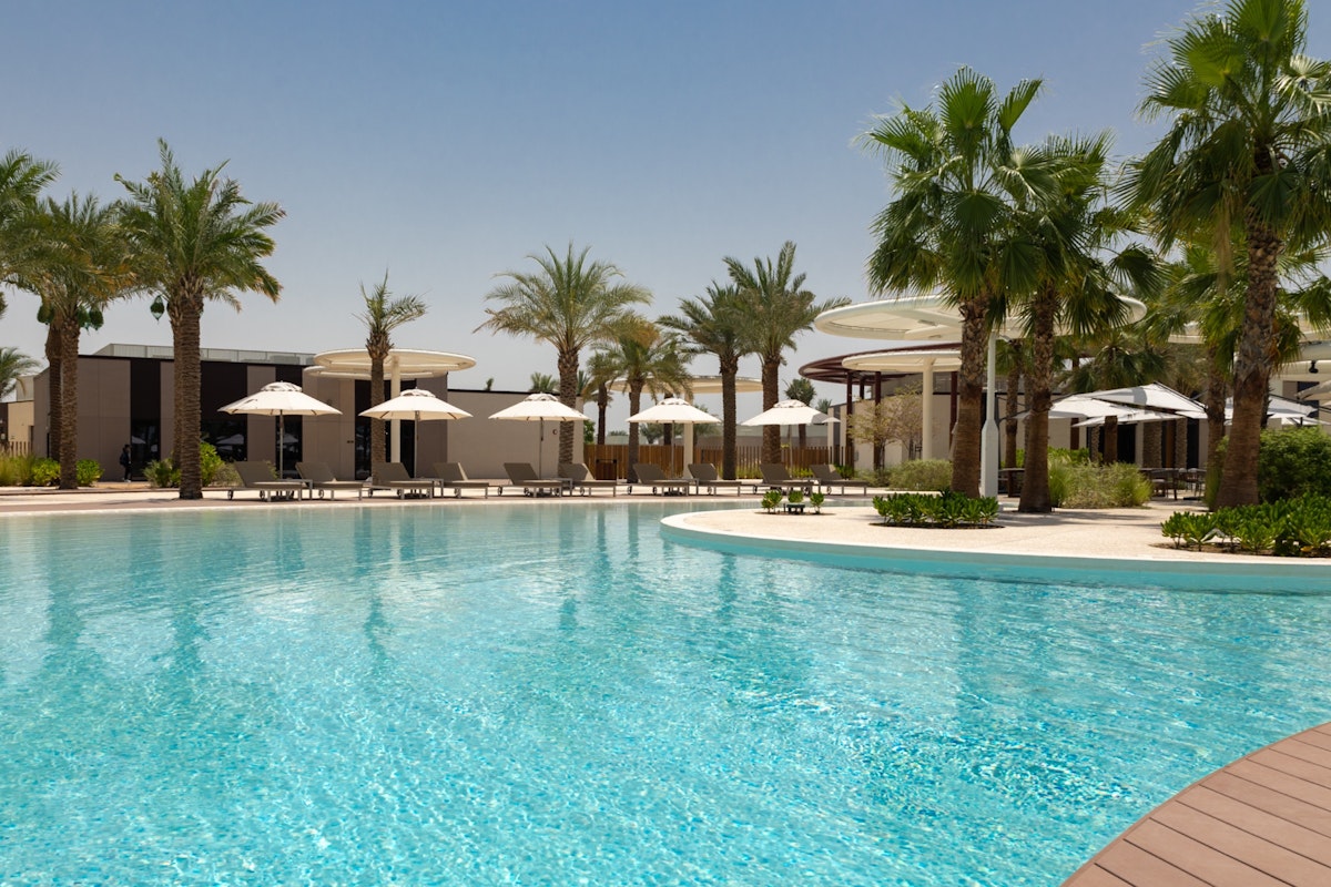 Swimming pool surrounded by umbrellas and palm trees with a sunny sky in the background.