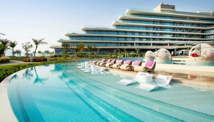 W Dubai The Palm A resort with a pool area featuring lounge chairs, surrounded by lush greenery, and a modern hotel building in the background.