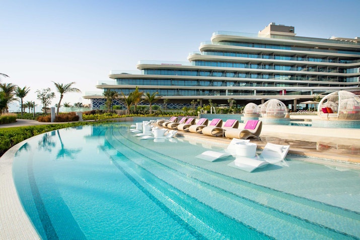 A resort with a pool area featuring lounge chairs, surrounded by lush greenery, and a modern hotel building in the background.