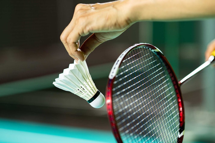 A close-up of a badminton racket with a shuttlecock being held by a hand.
