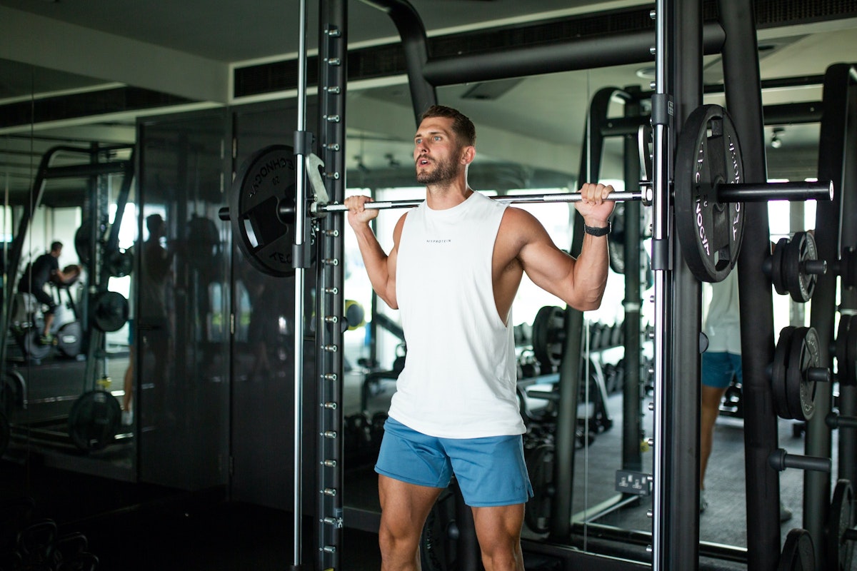 Use Privilee to reach your fitness goals A man performing a barbell shoulder press in a gym with various strength training equipment in the background.