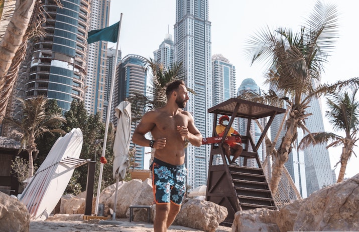 A man holding a beach lifeguard float, standing in front of a lifeguard tower with tall skyscrapers in the background.