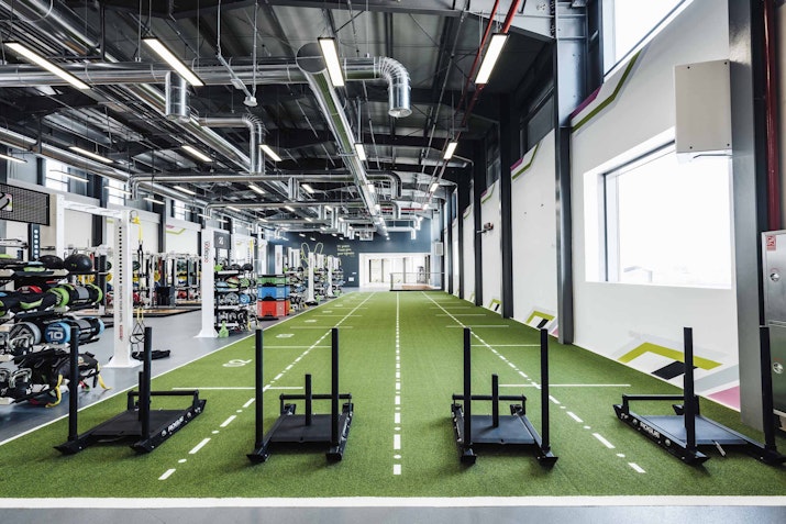 A gym with sled push equipment lined up on a turf track, with various workout stations visible in the background.