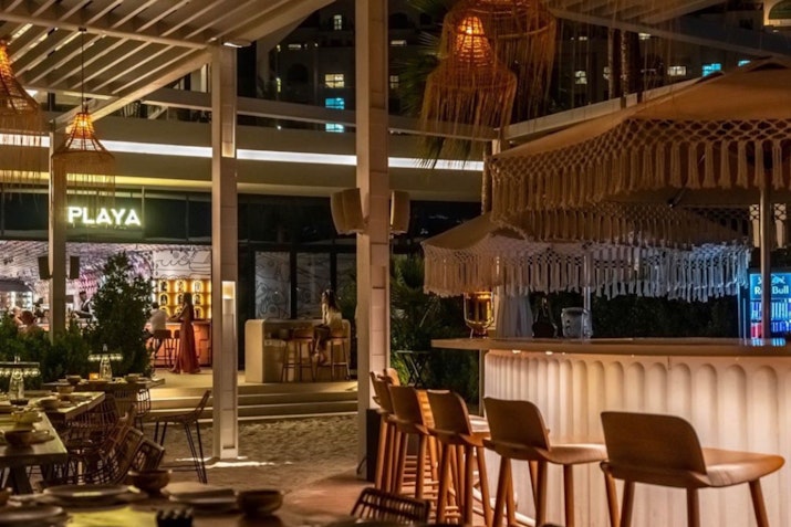 Chic outdoor bar with woven overhead lanterns and seating, illuminated with warm lights for a cozy and inviting atmosphere.