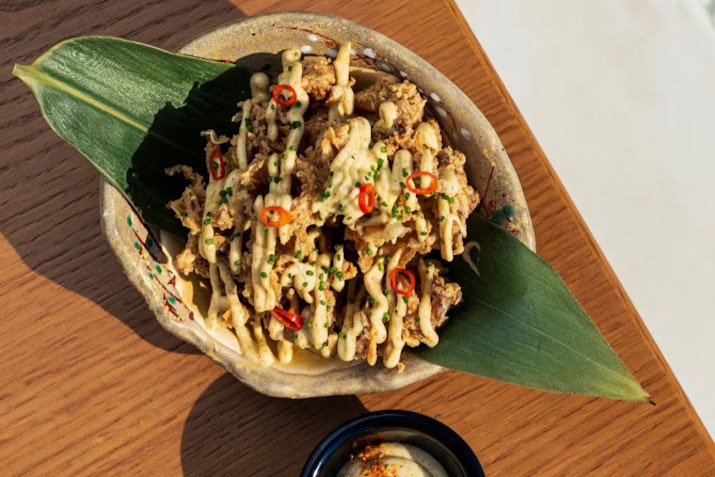 Close-up of a beautifully plated dish with crispy ingredients drizzled with creamy sauce and garnished with sliced red chilies and chives, presented on a decorative leaf.