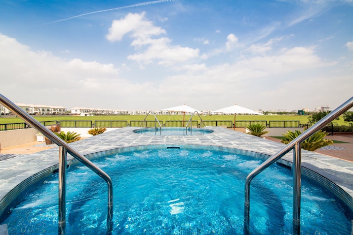 A hot tub with metal handrails and clear blue water, overlooking a green landscape with umbrellas in the distance.