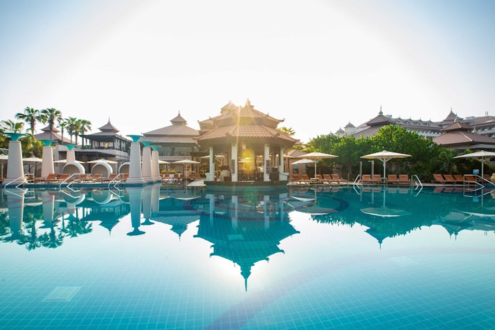 A serene pool with reflections of a pagoda-like structure and surrounding lounge chairs, bathed in sunlight.