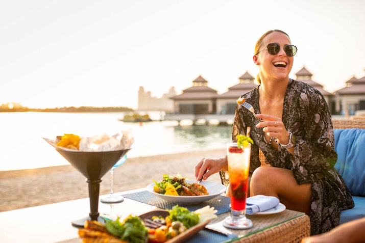 Woman enjoying a meal and drink by the beach with a smile.