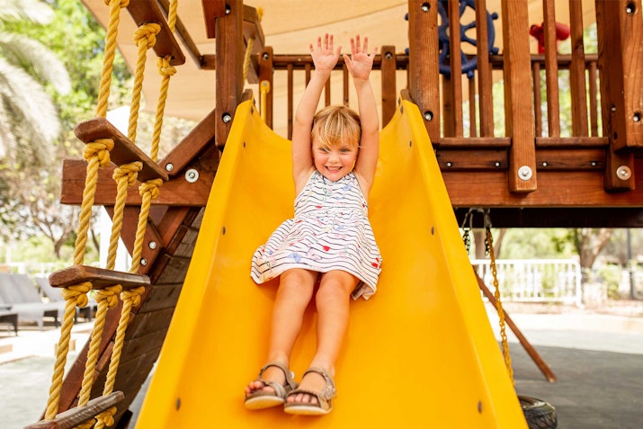 A child sliding down a yellow slide with arms raised, smiling.