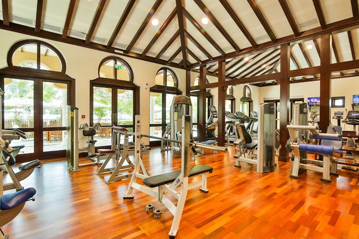 A well-equipped gym with wooden flooring and a high vaulted ceiling, featuring various fitness equipment and large windows.