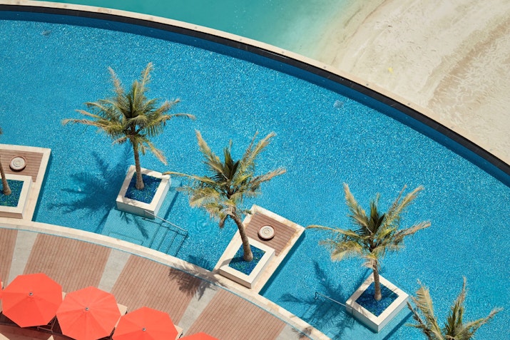 Top view of a pool with palm trees in pots and red umbrellas beside the water.