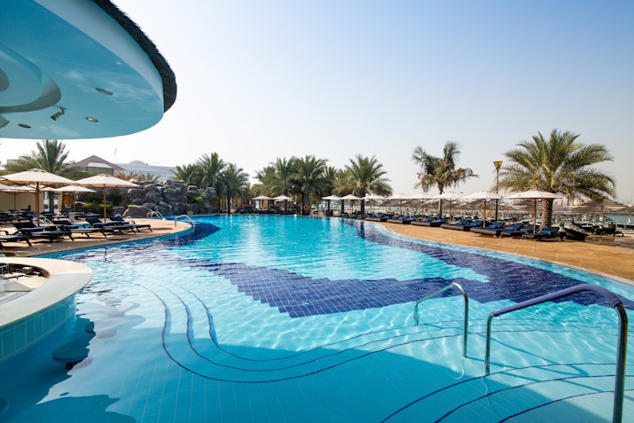 A swimming pool with a blue tile design and steps leading into the water, surrounded by palm trees and sun loungers.