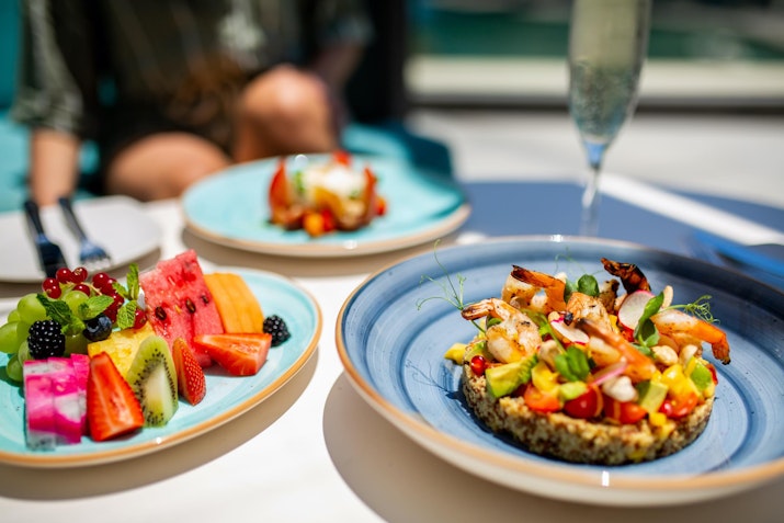 Plates of fresh fruit and a quinoa salad with shrimp and vegetables, with a glass of champagne in the background.