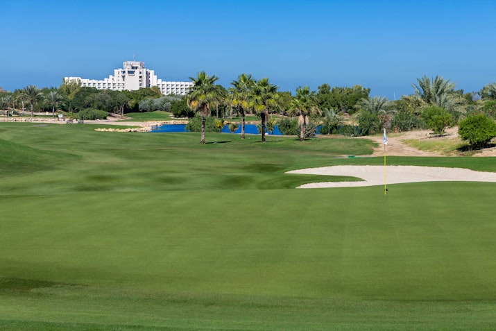 JA Beach Resort A scenic view of a golf course with lush green grass, palm trees, and a hotel in the background against a clear blue sky