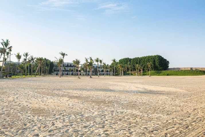 Open sandy area with palm trees and buildings in the background.