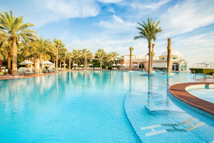 A serene swimming pool surrounded by palm trees and lounge chairs, with a beautiful sky in the background.