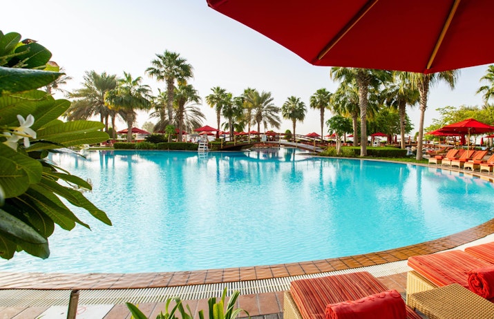 A relaxing poolside scene with red umbrellas, lounge chairs, and palm trees around the pool.