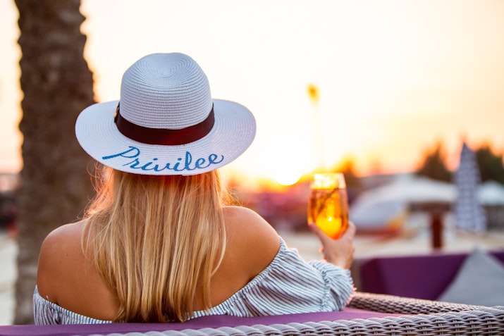 Woman with a 'Privilee' hat enjoying a drink during sunset.