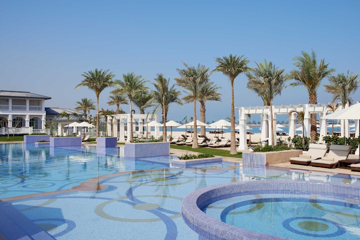 Luxury pool with circular patterns, surrounded by palm trees, lounge chairs, and a scenic view of the beach.