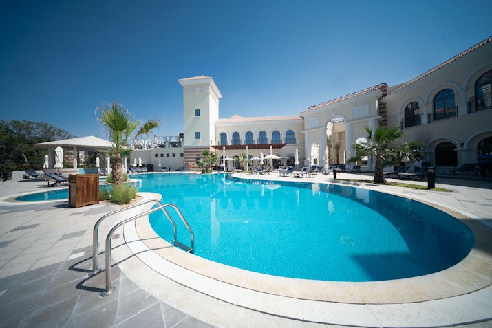 A tranquil swimming pool area at a resort, featuring a clear blue pool with a curved edge, surrounded by palm trees and sun loungers, and an elegant building in the background under a clear blue sky.