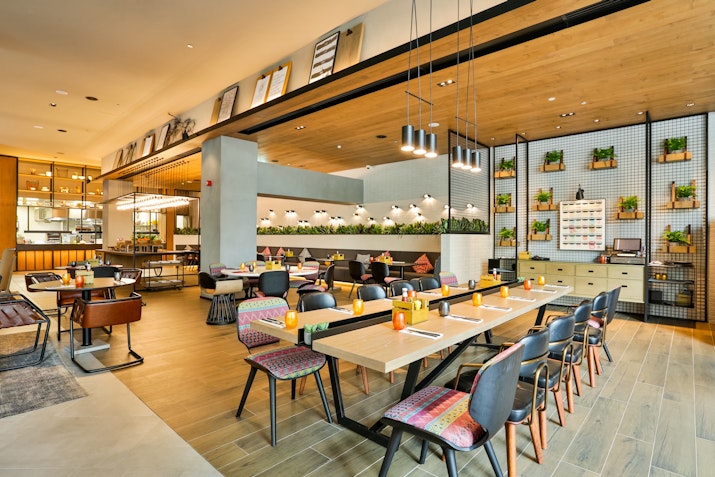 A modern restaurant interior with multiple dining tables, chairs, and plants decorating the space, creating a fresh and inviting atmosphere.