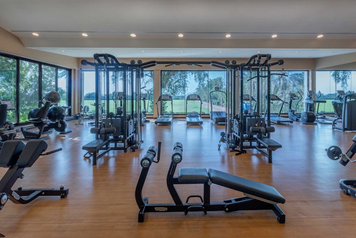 A well-lit gym with large windows offering a scenic view, featuring a variety of workout equipment including weights, benches, and machines for strength training.