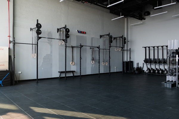 Minimalist gym space featuring gymnastic rings, pull-up bars, and a weight rack with barbells, set against a light gray wall on a spacious, dark-tiled floor.