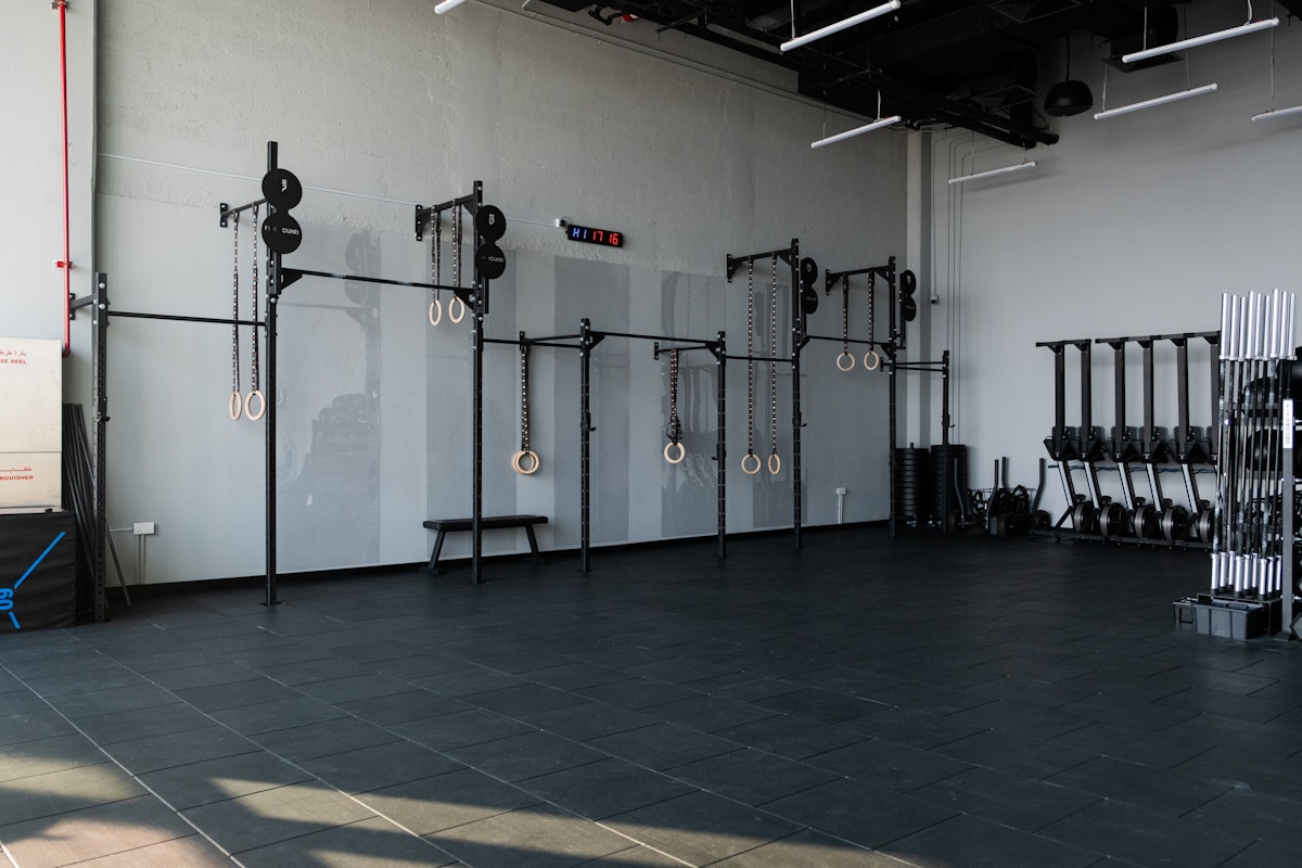 Minimalist gym space featuring gymnastic rings, pull-up bars, and a weight rack with barbells, set against a light gray wall on a spacious, dark-tiled floor.