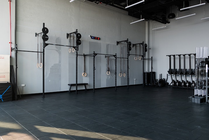 Minimalist gym space featuring gymnastic rings, pull-up bars, and a weight rack with barbells, set against a light gray wall on a spacious, dark-tiled floor.